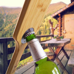 On a rustic porch with a mountainous landscape, trees, and a cabin, the BBQ Grill Scraper with Bottle Opener from Totally Bamboo is being used to open a beer bottle. The cozy outdoor setting is ideal for gathering around the grill.