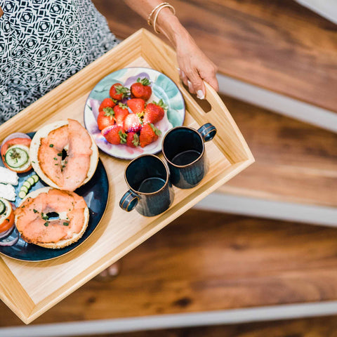 A person carries a Butler's Serving Tray with Handles by Totally Bamboo, elegantly adorned with delicious treats. The tray elegantly holds two bagels topped with smoked salmon and cream cheese, a dish of fresh strawberries, slices of cucumber and tomato, and two black mugs. The individual is stylishly dressed in a patterned top.