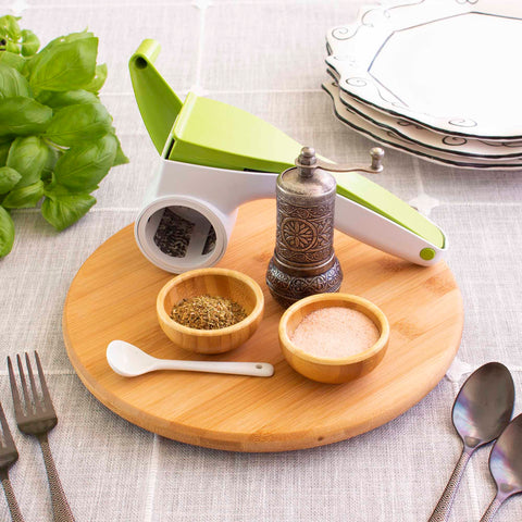 The TB Home® 10" Lazy Susan Turntable by Totally Bamboo elegantly displays a cheese grater, a pepper mill, small bowls of spices, a white spoon, and cutlery. Fresh basil and stacked plates enhance the background on a gray tablecloth, showcasing ideal kitchen organization.