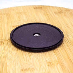 A round, black silicon pad rests on a light brown wooden surface, highlighting the grain and natural pattern. At its center, a small metal component allows for 360-degree rotation, making it an ideal addition to any kitchen organization setup like the TB Home® 14" Lazy Susan Turntable by Totally Bamboo.