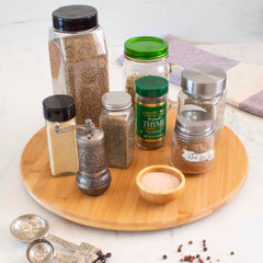 The TB Home® 14" Lazy Susan Turntable by Totally Bamboo elegantly displays an assortment of spice jars and shakers on its rotating surface, enhancing kitchen organization. A small bowl is filled with pink seasoning, while two ornate metal spoons featuring grape motifs complete the scene.