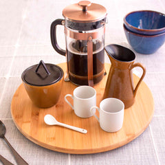 A French press with coffee is placed on a TB Home® 14" Lazy Susan Turntable by Totally Bamboo, accompanied by a small brown pitcher, two white cups, a brown lidded sugar bowl, and a white spoon. The turntable's 360-degree rotation enhances kitchen organization. In the background, two blue bowls rest on a light-colored tablecloth.