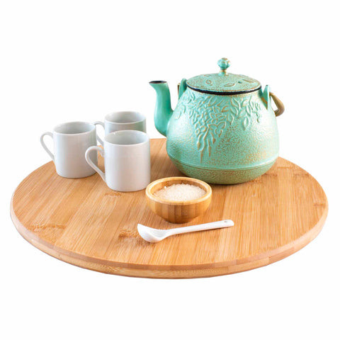 A green teapot with a floral design rests on the Totally Bamboo TB Home® 14" Lazy Susan Turntable, providing effortless 360-degree rotation. Nearby are three white mugs, a small wooden bowl filled with sugar, and a white spoon. This arrangement creates an inviting and well-organized tea setting.