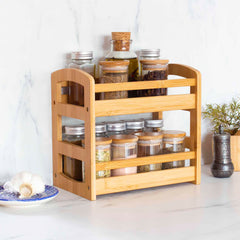 A Totally Bamboo 2-Tier Spice Caddy, crafted from bamboo, is displayed on a marble countertop holding an assortment of spice and oil jars. Next to it sits a garlic bulb on a small blue and white plate, complemented by a small potted plant against the backdrop of a sleek white marble wall.