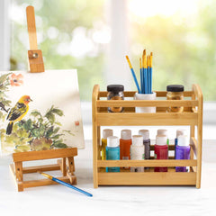 Displayed on a small easel is a painting of a bird with flowers, alongside the Totally Bamboo 2-Tier Spice Caddy, which is creatively used to organize various paint bottles, jars, and brushes. A brush lies on the table while the background reveals a window featuring softly blurred greenery.