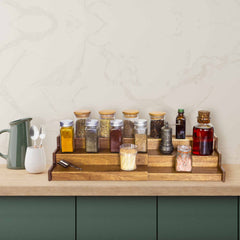 On the kitchen counter, a Totally Bamboo TB Home® 3-Tier Expandable Spice Rack holds various jars of spices and oils. A green ceramic pitcher with utensils is positioned on the left. The counter and bottom cabinets are a muted green, set against a light marbled wall.