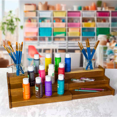 A wooden display, reminiscent of a TB Home® 3-Tier Expandable Spice Rack by Totally Bamboo, holds colorful paint bottles in neat rows. Jars of paintbrushes flank the sides, while shelves filled with art supplies add a vibrant touch to the scene, much like an artist's lively spice storage solution.