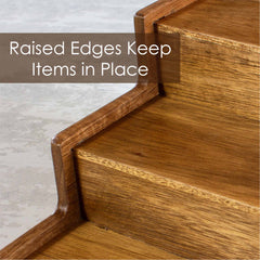 A close-up of wooden steps with raised edges showcases the TB Home® 3-Tier Expandable Spice Rack by Totally Bamboo. Text overlay states, "Raised Edges Keep Items in Place." The light brown wood design ensures secure spice storage by preventing items from falling off.