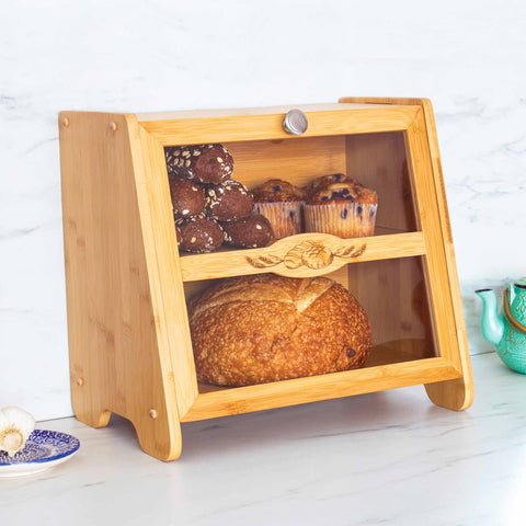 The Farmhouse Bread Box by Totally Bamboo, featuring a stylish glass door, stores a loaf of bread on the bottom shelf and an array of muffins on the top. Placed on a marble countertop, it exudes rustic charm and is complemented by the presence of a garlic bulb and a small turquoise teapot nearby.