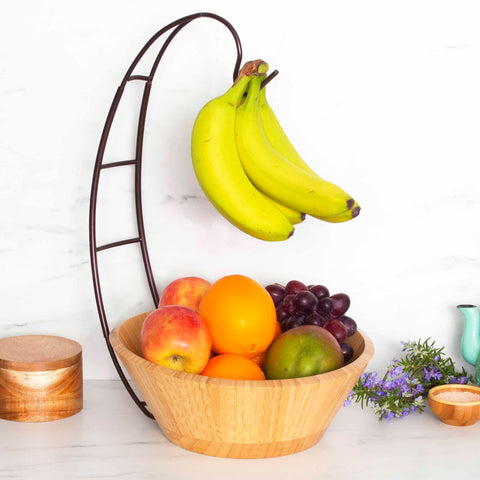 A Totally Bamboo Fruit Bowl with a Banana Holder sits on a sleek white marble countertop, filled with apples, oranges, a pear, and purple grapes. Above the bowl, bananas hang elegantly from the banana holder. Nearby, a small vase with flowers and two wooden containers complete the charming scene.