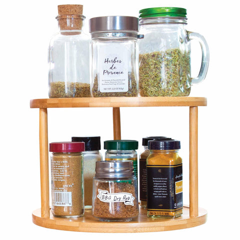 The TB Home® 2-Tier 10" Lazy Susan Turntable by Totally Bamboo beautifully showcases a collection of spice jars. The top tier holds larger glass jars with cork and metal lids, while the bottom features smaller plastic and glass containers, each carefully labeled with various spices.