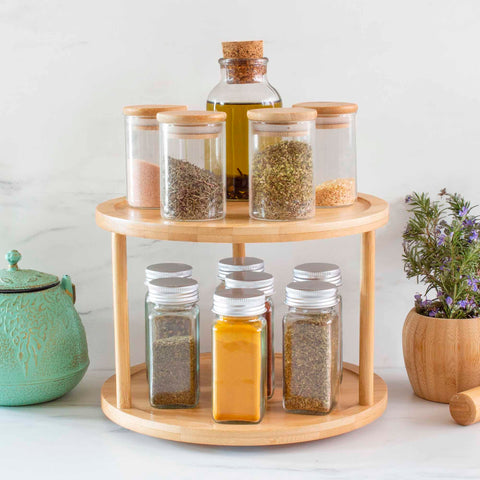 A TB Home® 2-Tier 10" Lazy Susan Turntable by Totally Bamboo elegantly organizes various spice jars. The top tier showcases jars with cork and wooden lids, while the bottom tier contains jars with metal lids. A ceramic teapot and a potted plant add charm to this captivating setup.