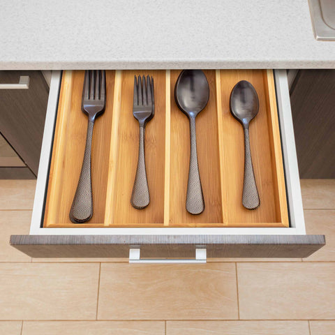 As you open the kitchen drawer, the Totally Bamboo Flatware Drawer Organizer comes into view, neatly arranging four pieces of flatware: a dinner fork, salad fork, tablespoon, and teaspoon. Each piece features textured handles. The elegant contrast between the wooden floor and light countertop enhances the kitchen's sophisticated aesthetic.