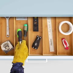 A person wearing a yellow glove arranges a drawer using the Totally Bamboo Expandable Drawer Organizer, neatly placing tools such as a wrench and screwdrivers. This wooden organizer includes precise bamboo dividers for seamless kitchen organization and efficient tool storage.