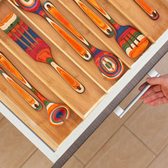 Someone opens a kitchen drawer revealing a Totally Bamboo Expandable Drawer Organizer, featuring neatly arranged handcrafted wooden utensils. The utensils, decorated with detailed orange, blue, and green patterns, fit perfectly among the bamboo dividers. A hand can be seen pulling the drawer handle.