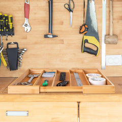 An Expandable Drawer Organizer by Totally Bamboo transforms the wooden workbench into a hub of organized precision. Dividers ensure tools like screwdrivers, rulers, tape measures, and levels are neatly arranged on a tray. Above the workspace, wrenches, screwdrivers, scissors, and a saw hang methodically on the wall. A hammer is positioned conveniently on the right side. This setup perfectly blends form and function!