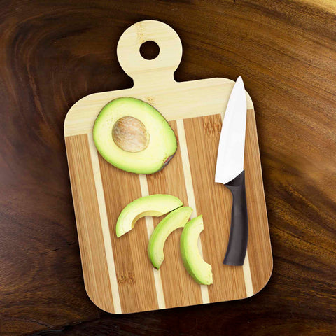 Two-tone striped bamboo paddle board with avocado and paring knife on dark brown wooden table