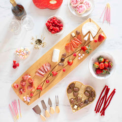 A Del Mar Extra-Large Charcuterie Board by Totally Bamboo, crafted from Moso bamboo and measuring 30" x 8-1/2", elegantly showcases an assortment of cheeses, meats, strawberries, and sweets arranged with artistic flair. Surrounding this sophisticated display on a white marble surface are raspberries, chocolate boxes, bottles of wine, and strawberry-themed desserts.