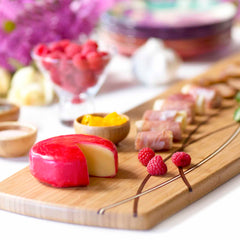 The Del Mar Extra-Large Charcuterie Board by Totally Bamboo, measuring 30" x 8-1/2", elegantly showcases a delightful assortment of appetizers: a partially sliced round of red cheese, fresh raspberries, bowls filled with mango chutney, and savory prosciutto-wrapped treats. In the background, a glass bowl overflows with raspberries beside blurred floral accents.