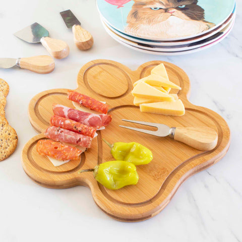 Sliced meats, cheese cubes, and peppers are artfully arranged on a Totally Bamboo 11" x 10" paw-shaped cutting board. This board offers a knife-friendly surface and comes with a small wooden-handled knife. Nearby, there's a stack of plates featuring cat designs with some crackers partially visible in the scene.