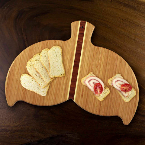 A Totally Bamboo Whale Tail Shaped Cutting Board, measuring 14-1/2" x 10-1/2", showcases several slices of bread on one side and two open-face sandwiches topped with sliced meat and cherry tomatoes on the other. This charming board, resting on a dark wooden table, can also serve as delightful kitchen wall art.