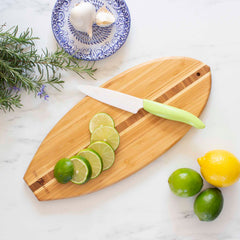 The 14-1/2" x 6" Li'l Surfer Cutting Board from Totally Bamboo comes with a lime-green-handled knife perfect for slicing limes. Alongside, you'll find whole limes and a lemon resting nearby. A small blue and white dish holds garlic and ginger, while fresh rosemary is elegantly arranged on the white marble surface.