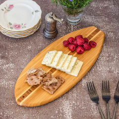 On the Li'l Surfer Cutting Board by Totally Bamboo, sliced cheese, raspberries, and crispbread are displayed. The scene includes three forks, stacked floral plates, a small ornate pepper grinder, and a glass vase with blue flowers resting on a textured surface nearby.