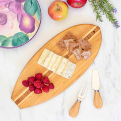 The Li'l Surfer Cutting Board by Totally Bamboo, measuring 14-1/2" x 6", elegantly showcases sliced cheese, raspberries, and crackers. Next to it, two cheese knives are ready for use. The scene on the marble surface is completed with apples, a vibrant plate, and rosemary sprigs surrounding the setup.