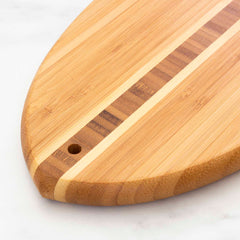 A close-up of the Totally Bamboo Li'l Surfer Cutting Board, measuring 14-1/2" x 6", highlights its distinctive surfboard-shaped design with a stylish two-toned stripe running down the center. It includes a convenient small hole near the edge for easy hanging. Displayed against a light marble surface, the rich wood grain is prominently visible, adding to its elegant appeal.