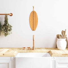 A modern kitchen featuring wooden countertops, a white farmhouse sink, and a copper faucet. A Totally Bamboo Li'l Surfer Cutting Board (14-1/2" x 6") is displayed on the white wall, while herbs dangle to the left. A white vase on the right contains wooden utensils, enhancing the coastal charm.