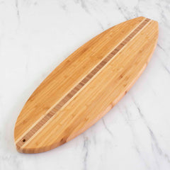 The Totally Bamboo "Surfboard Shaped Cutting Board" (23" x 7-1/2") with a central striped detail, designed from bamboo for a knife-friendly surface, is elegantly displayed on a white marble surface.