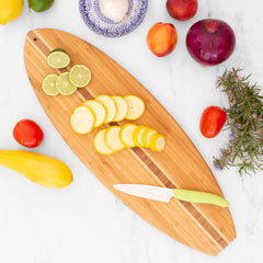 A top view showcases the Totally Bamboo Surfboard Shaped Cutting Board, measuring 23" x 7-1/2", adorned with sliced yellow squash and limes. A green-handled knife complements the display, surrounded by whole vegetables and herbs: onion, tomatoes, rosemary, and peaches—all elegantly arranged on a marble surface.