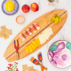 A Surfboard Shaped Cutting Board by Totally Bamboo, measuring 23" x 7-1/2", is adorned with stacked cheese slices, prosciutto-wrapped cheese, and crackers. The surrounding setup includes peaches, a lemon, a salt dish, pepperoncini, and a floral plate with vibrant utensils. Small red flowers enhance the charm of this knife-friendly serving surface.