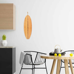 This minimalist kitchen showcases the Totally Bamboo Surfboard Shaped Cutting Board, measuring 23" x 7-1/2", displayed on the white wall. It complements the wooden table accompanied by two black chairs and a teapot. Three lemons add a pop of color, and a gray cloth introduces texture to the scene. A plant is placed on the dark countertop for a natural touch.