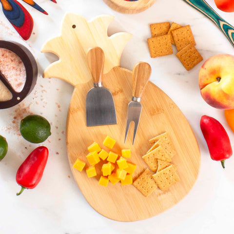 A Totally Bamboo Pineapple Shaped Cutting Board, measuring 14-3/8" x 7-1/2", elegantly displays cheese cubes, crackers, a cheese knife, and fork. The charcuterie is artistically surrounded by whole limes, peaches, red peppers, and a salt container on a pristine white surface.