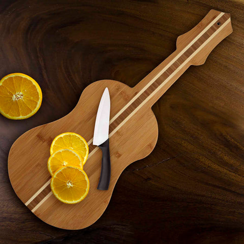 The Totally Bamboo Ukulele Shaped Cutting Board, measuring 22" x 9", captures a guitar-like design and displays three orange slices alongside a knife on a dark wooden surface. In the upper left corner, two extra orange halves complement its functionality with an artistic flair, making it ideal as both a practical kitchen tool and decorative wall art.