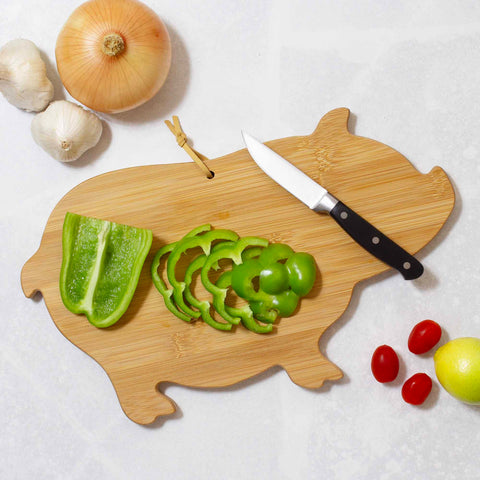 A Totally Bamboo Pig Shaped Cutting Board, measuring 15-5/8" x 9-1/2", showcases sliced green bell pepper with a knife, while an onion, garlic bulbs, cherry tomatoes, and a lemon complement this delightful gift cutting board on the light countertop.