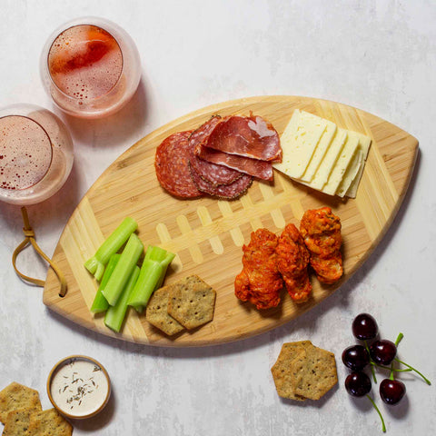 On a Totally Bamboo Football Shaped Cutting Board, measuring 15" x 8-1/2", there is an assortment of sliced meats, cheese, buffalo chicken wings, celery sticks, and crackers. Nearby are two glasses of a red beverage, a small bowl of dip, and cherries for snacking.