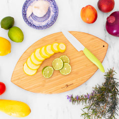 The Totally Bamboo Football Shaped Cutting Board, measuring 15" x 8-1/2", features an arrangement of sliced yellow squash and lime alongside a green-handled knife. Nearby ingredients like garlic, peach, onion, lime, lemon, and zucchini rest on the marble surface. This colorful display is complemented by a blue-patterned plate, making it an ideal choice for a tailgate serving board.