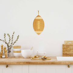 A minimalist kitchen interior features a bamboo cutting surface complemented by bowls, a pepper mill, and a vase with leafy branches. Above, the Football Shaped Cutting Board by Totally Bamboo, measuring 15" x 8-1/2", is elegantly displayed on the white wall, adding charm to the serene space.