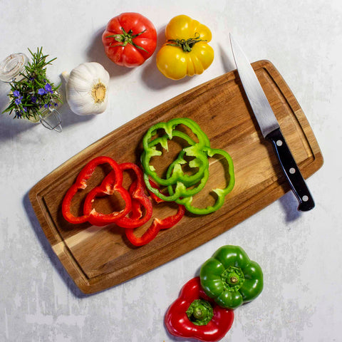 The TB Home® Cutting Board with Juice Groove by Totally Bamboo, measuring 14" x 7", displays sliced red and green bell peppers along with a knife and whole peppers. Nearby are a red and yellow heirloom tomato, a bulb of garlic, and a sprig of rosemary on a light textured surface.