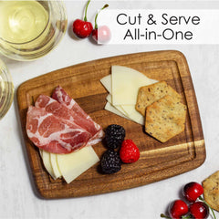 An elegant array of assorted meats, cheese slices, crackers, and berries are beautifully displayed on a Totally Bamboo TB Home® Small Cutting Board with Juice Groove, crafted from rich acacia wood. Nearby sits a glass of white wine and a serving of cherries. In the top right corner, the text "Cut & Serve All-in-One" emphasizes its convenient juice groove feature.