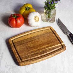 A Totally Bamboo TB Home® Small Cutting Board with Juice Groove, measuring 8" x 6", crafted from acacia wood, sits on the kitchen counter next to a knife. Beside it are a red and yellow tomato, a bulb of garlic, and a jar of herbs. Its juice groove ensures no mess disrupts your culinary prep area.