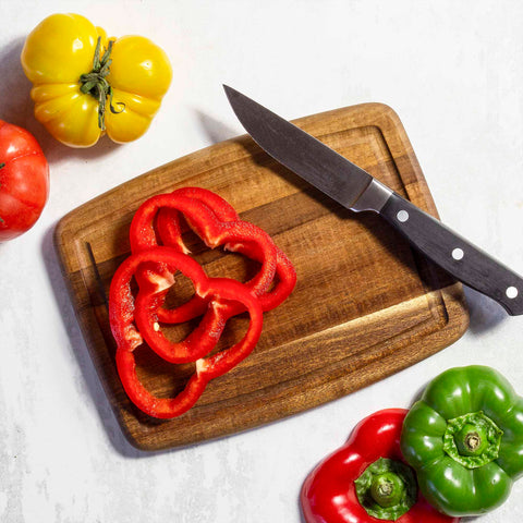 Slices of red bell pepper are placed on a Totally Bamboo TB Home® Small Cutting Board with Juice Groove, 8" x 6", alongside a knife. Surrounding the acacia wood board are whole yellow, green, and red bell peppers, along with a tomato on a white surface.