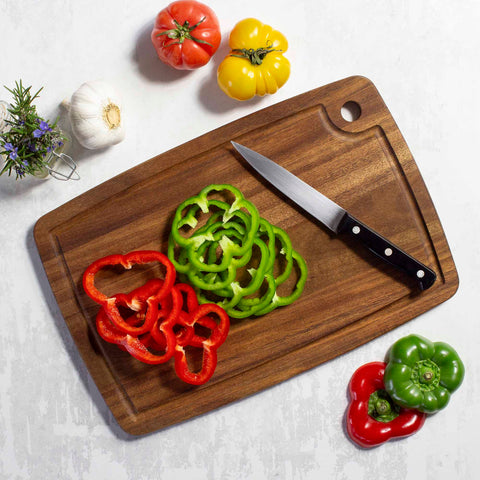 The TB Home® Cutting Board with Juice Groove by Totally Bamboo, measuring 15" x 10", showcases sliced red and green bell peppers accompanied by a knife. Surrounding the board are vibrant red, yellow, and green tomatoes, garlic cloves, and a sprig of rosemary on a light-textured surface.