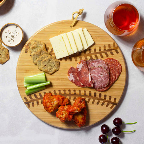 A baseball-shaped cutting board from Totally Bamboo, measuring 12" x 12", holds sliced cheese, salami, crackers, celery sticks, buffalo wings, and a glass of rosé. Nearby, cherries are scattered—perfect for game day entertaining.