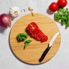 A basketball-shaped bamboo cutting board from Totally Bamboo, measuring 12" x 12", holds a knife and sliced tomatoes, making it perfect for game day entertaining. Nearby are basil leaves, a red onion, a bulb of garlic, whole tomatoes, and peppercorns on a light surface.