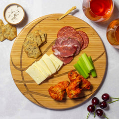 The Totally Bamboo Basketball Shaped Cutting Board, measuring 12" x 12", holds an array of sliced cheese, cured meats, celery sticks, and buffalo wings. Nearby are crackers and dipping sauce, making it perfect for game day entertaining. Glasses filled with a pink beverage and a few cherries complete the spread on a crisp white surface.