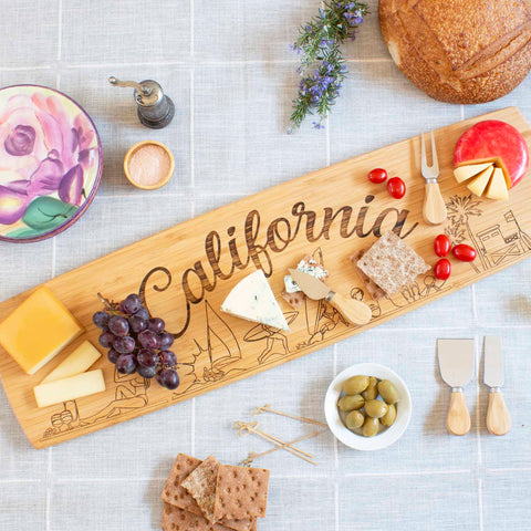 The Totally Bamboo California Engraved XL Charcuterie Board, measuring 30" x 8-1/2", elegantly displays a selection of cheeses, grapes, cherry tomatoes, crackers, and olives. Accompanying this board are a round loaf of bread, a floral plate, a small bowl of salt, and a pepper grinder atop a tablecloth.