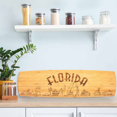 In a kitchen with a light blue wall, a shelf displays jars of pasta, grains, and spices. The Florida Engraved XL Charcuterie Board by Totally Bamboo, measuring 30" x 8-1/2", rests against the countertop alongside a leafy plant and spice jars, enhancing the Florida-inspired decor.